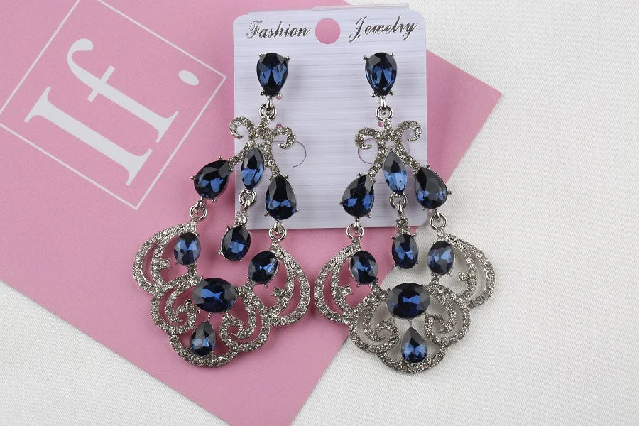 Un par de aretes colgantes elegantes con piedras azules y detalles plateados brillantes, exhibidos en un soporte de joyería sobre un fondo rosa.